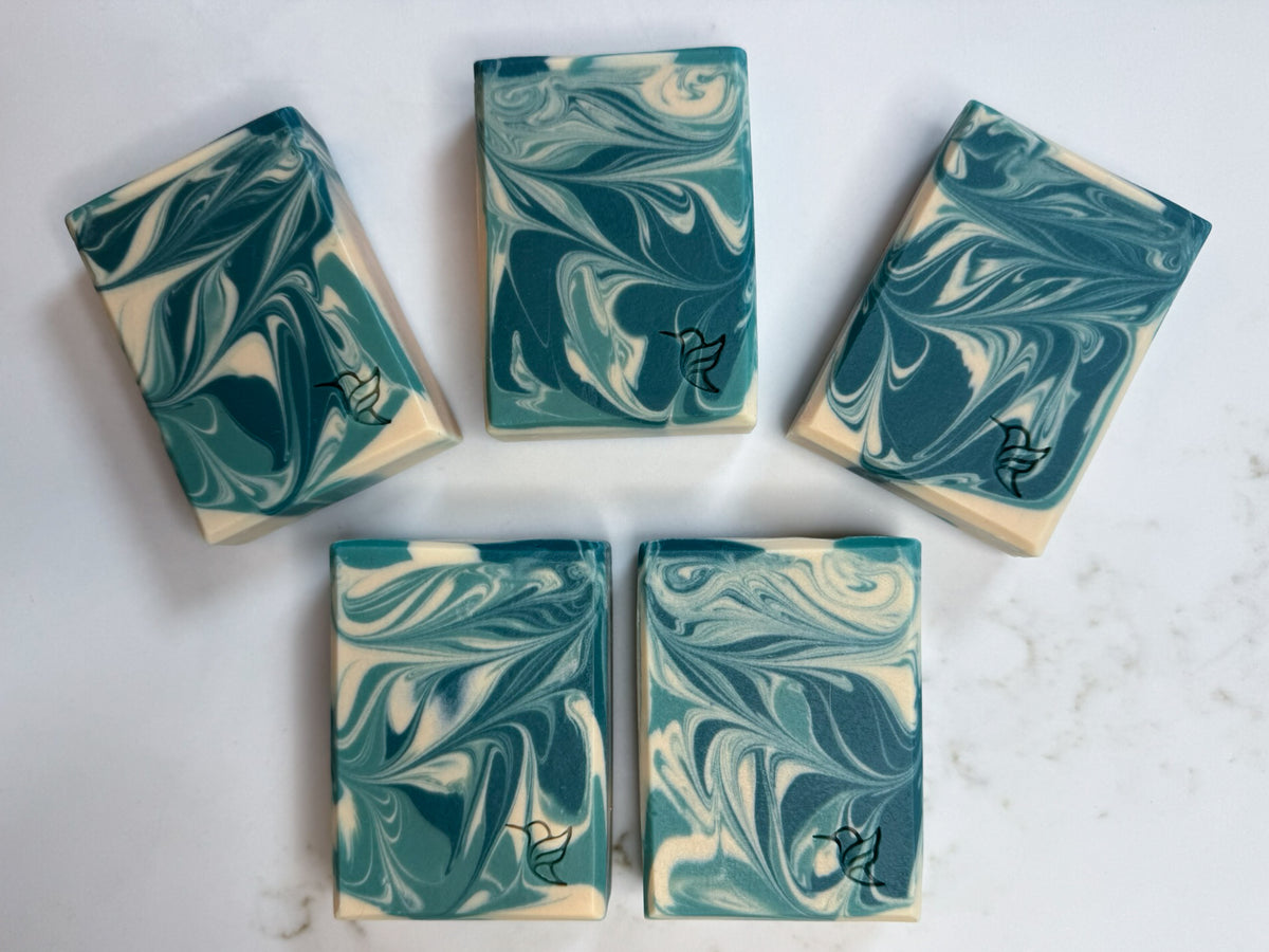 Five handmade Bay Leaf & Tobacco triple butters, tallow and goat milk soap bars displaying unique teal, green and ivory swirls with hummingbird maker's marks.
