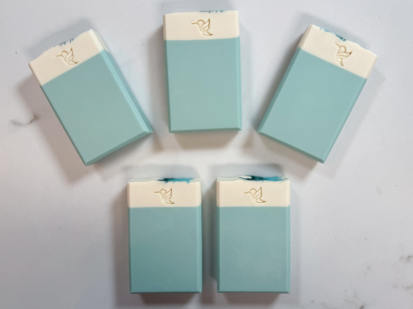 Five artisan cold process vegan soap bars with a teal and cream two-tone design and hummingbird logo stamp, displayed on a white quartz slab.