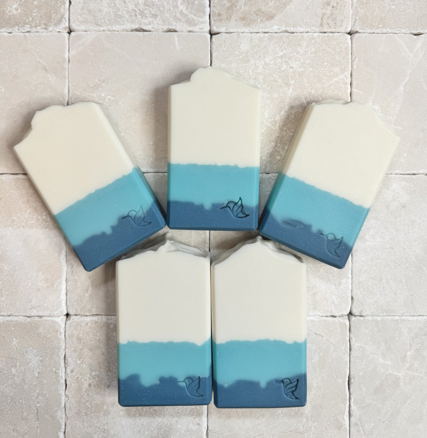 Five rectangular bars of Eucalyptus & Spearmint vegan triple butter soap by Happy Hummingbird Soapery, each featuring a white-to-blue color gradient and an embossed hummingbird logo, displayed on a light stone tile surface.