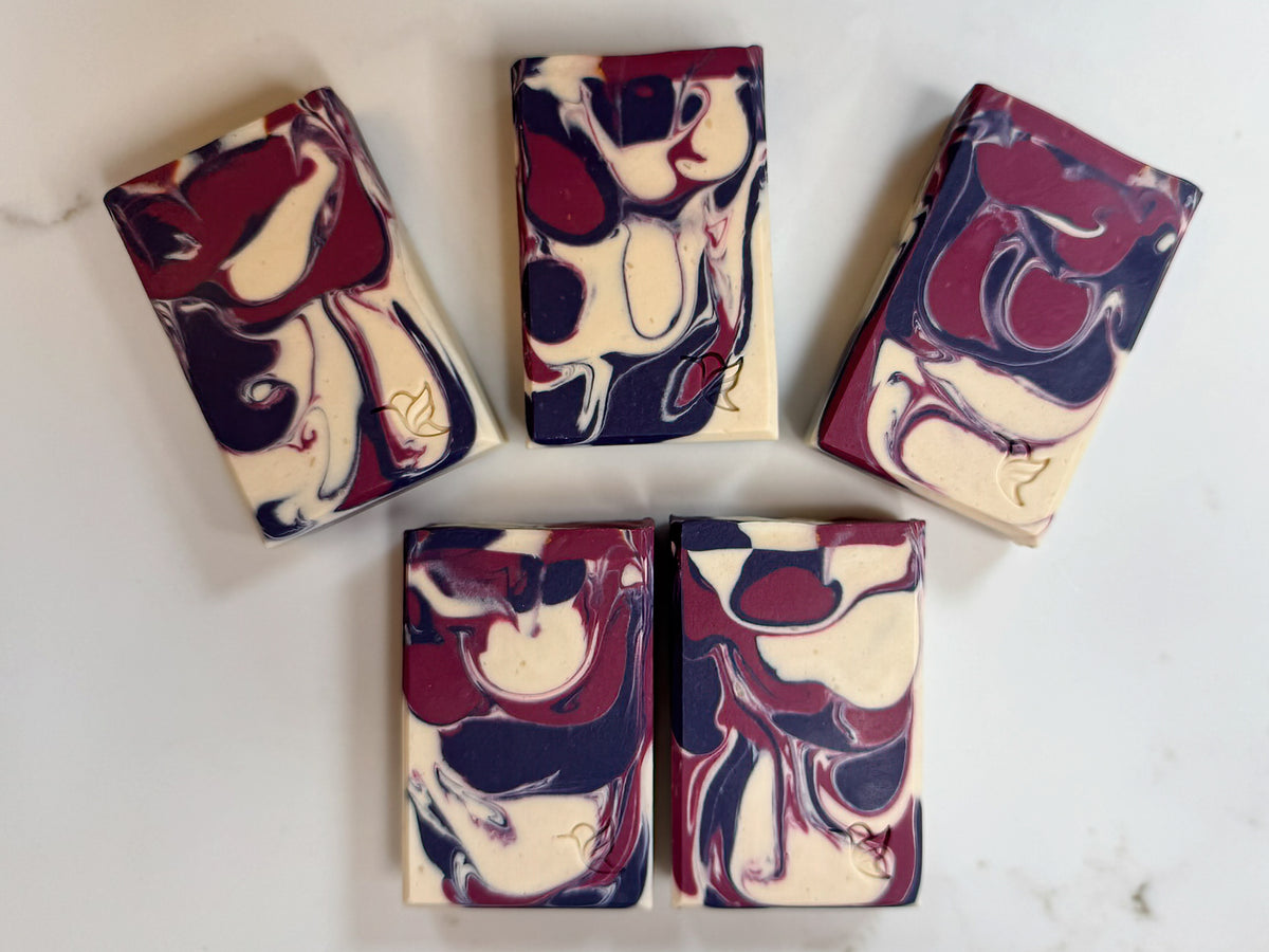 Five bars of Black Raspberry Vanilla vegan triple butter oat milk soap arranged on a quartz background, showing burgundy, deep navy and cream swirls with hummingbird maker’s marks.