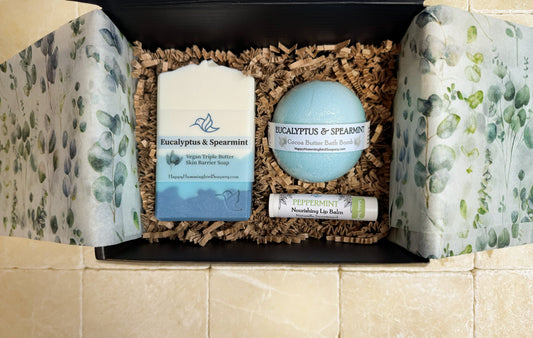 Happy Hummingbird Soapery Eucalyptus & Spearmint gift set in an open black box, featuring a round bath bomb, artisan soap bar, and peppermint nourishing lip balm, arranged on natural brown crinkle paper.