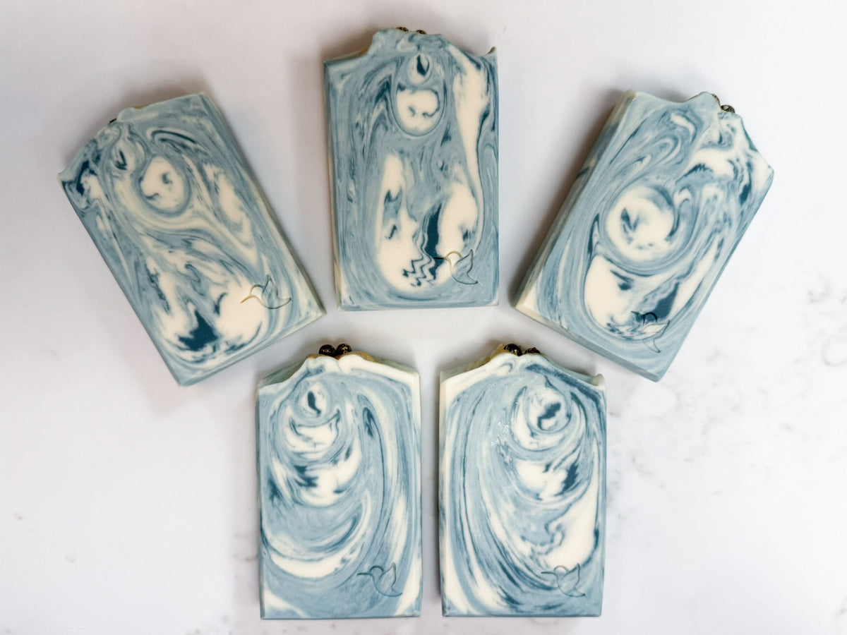 Five handcrafted Fraser Fir soap bars arranged in a fan on a white marble surface, displaying teal and cream swirls, stamped hummingbird logos, and textured tops with juniper berries.