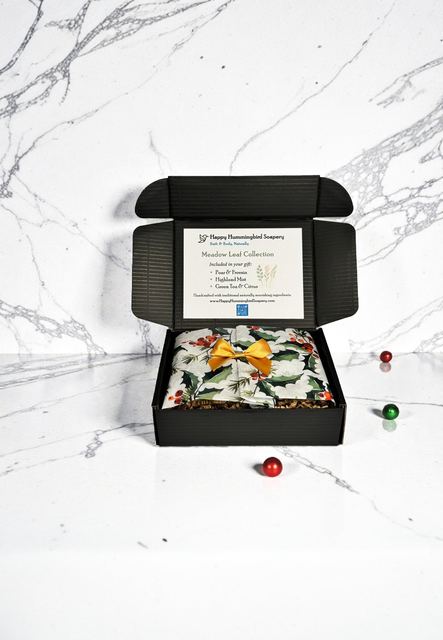 Open box with tissue folded and bow:
“Meadow Leaf Collection gift box with holiday tissue folded closed and gold satin bow — Happy Hummingbird Soapery.