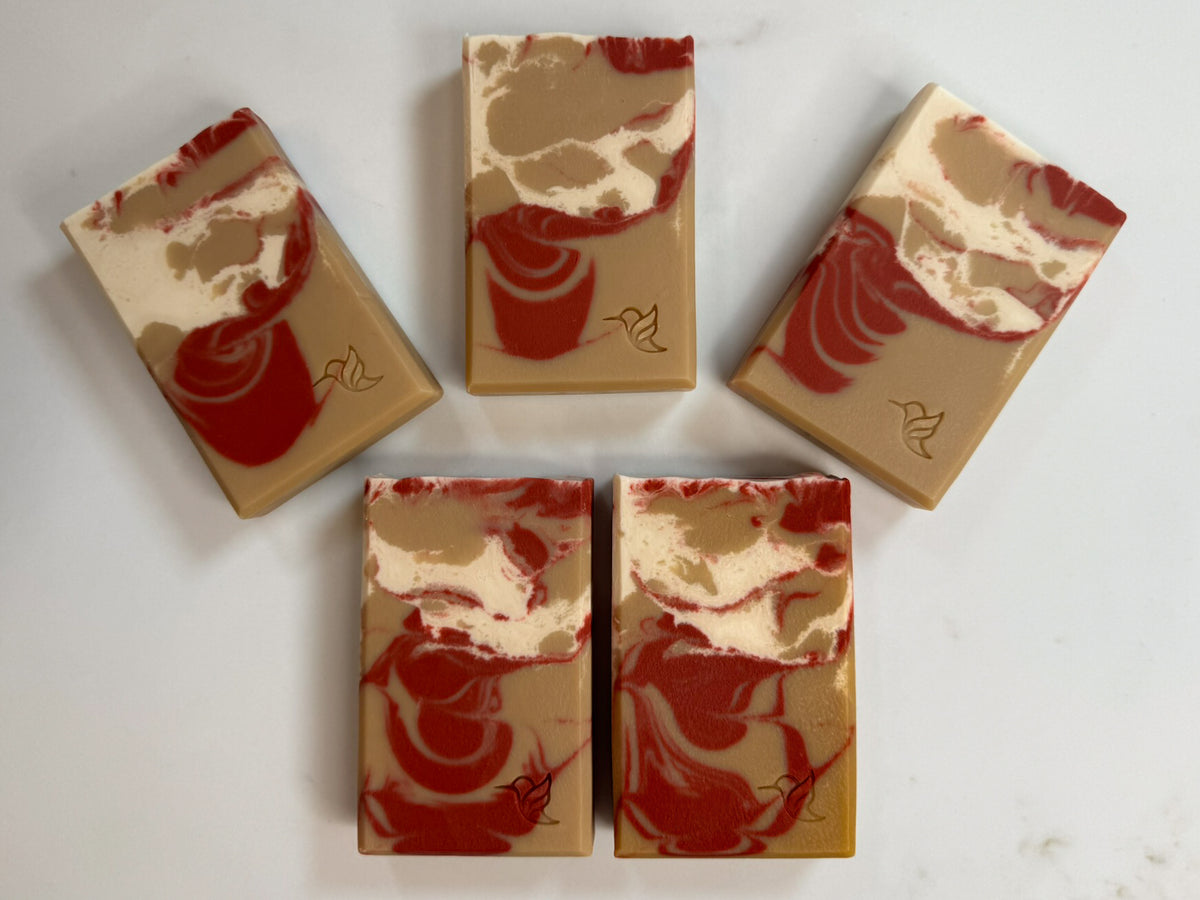 Five handcrafted bars of Oak Barrel Spiced Apple Cider cold process soap arranged in a fan on a white marble background, displaying the natural variations of the deep red, golden cider and cream oat milk marbled swirls and stamped hummingbird logos.