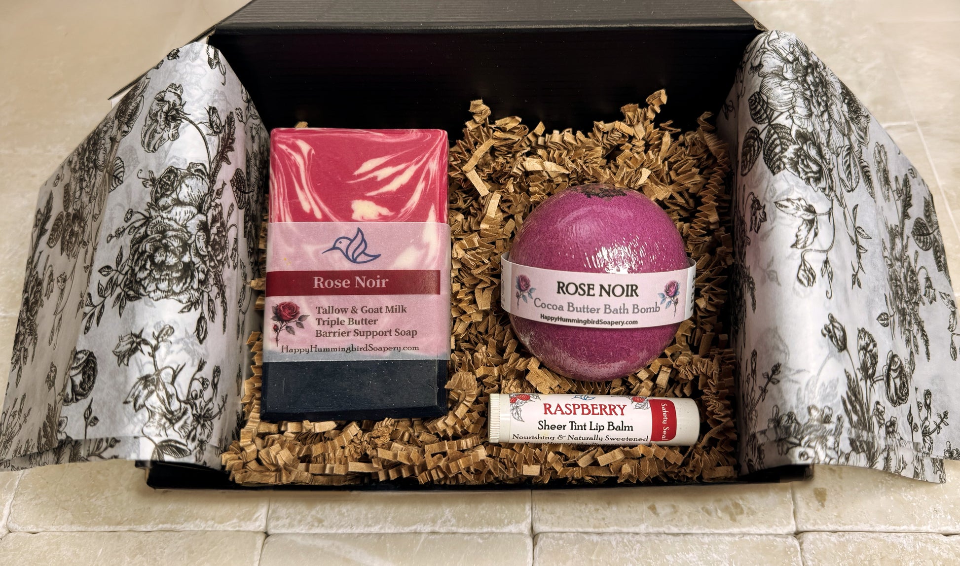 Happy Hummingbird Soapery Rose Noir gift set in an open black box, featuring a round bath bomb, artisan soap bar, and Sheer Tint Raspberry nourishing lip balm, arranged on recycled brown crinkle paper.