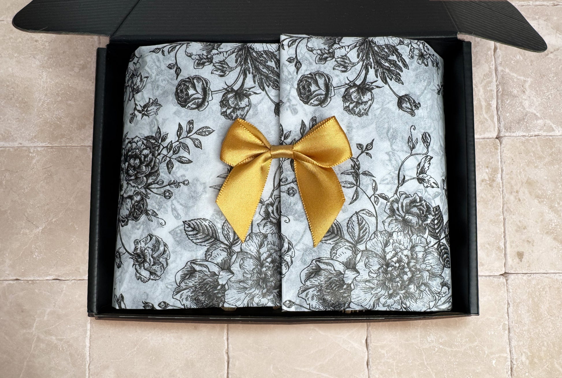 Rose Noir black premium gift box with floral tissue paper covering bath products, resting on a travertine stone surface.