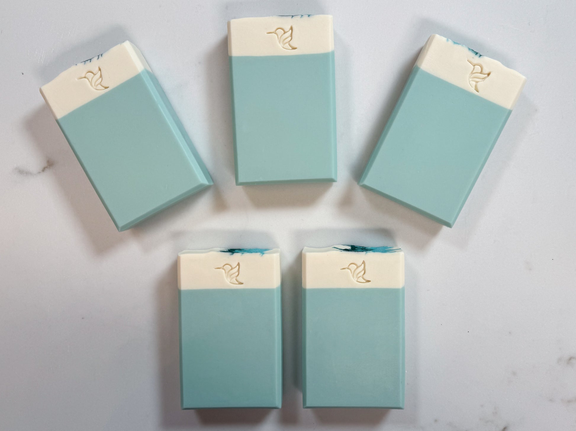 Five artisan cold process vegan soap bars with a teal and cream two-tone design and hummingbird logo stamp, displayed on a white quartz slab.