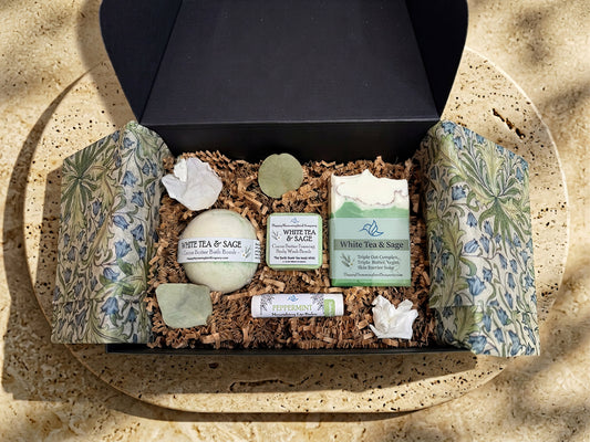 Happy Hummingbird Soapery White Tea & Sage gift set in an open black box, featuring a round bath bomb, foaming body wash bomb, artisan soap bar, and peppermint nourishing lip balm, arranged on natural brown crinkle paper.