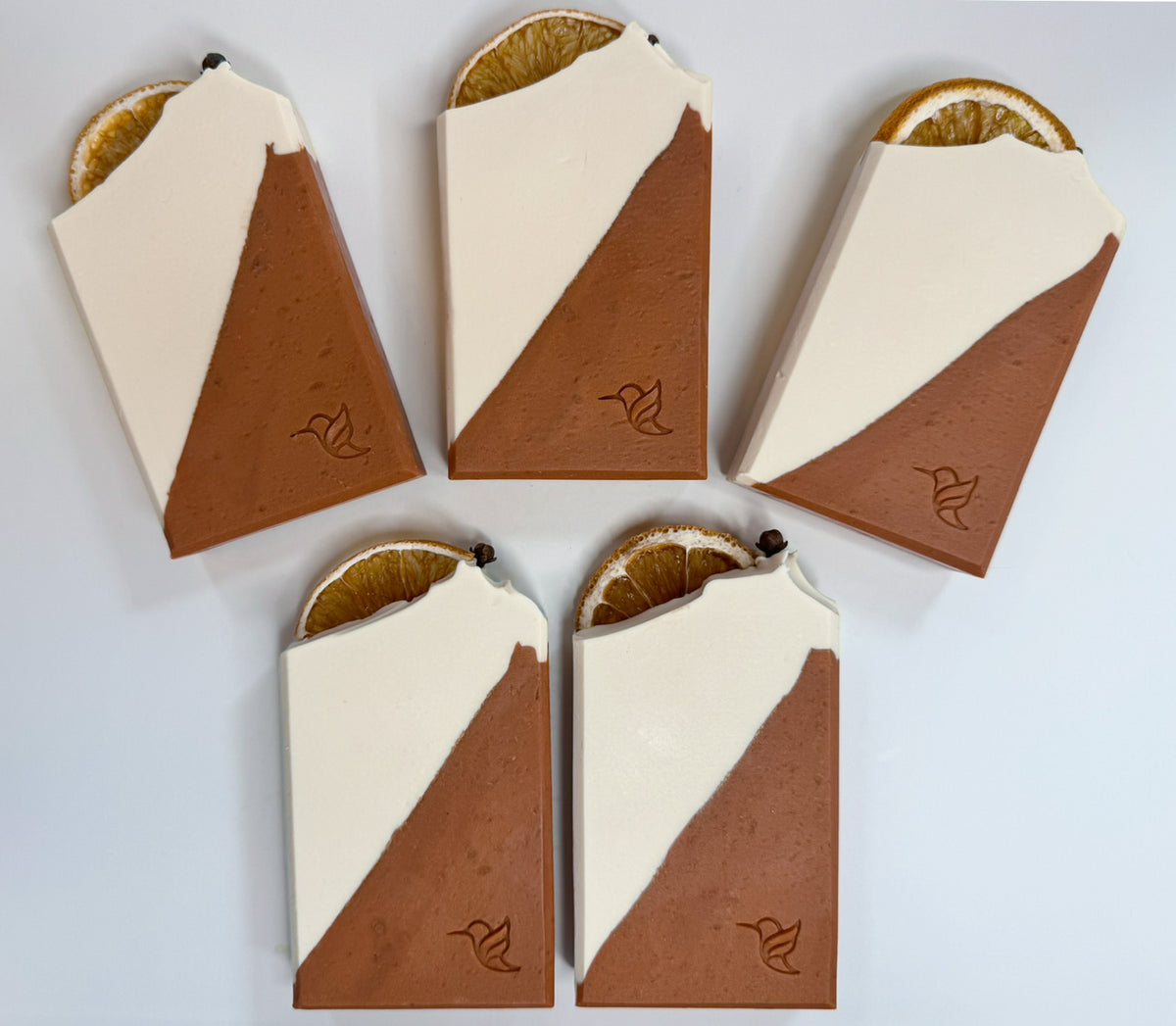 Five handcrafted Winter Orange and Clove triple butter, tallow and goat milk soap bars arranged in a fan on a white quartz background, displaying a cream and terracotta red clay diagonal design, stamped hummingbird logo, and dried orange slices and whole cloves.