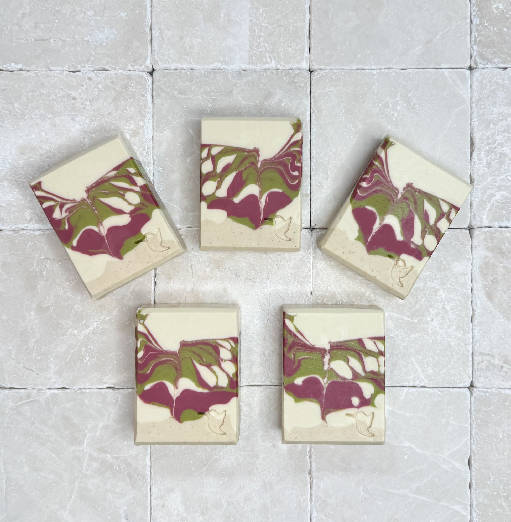 Five bars of Fig, Cassis & Oatmilk Soap by Happy Hummingbird Soapery, featuring a cream, green, and pink swirl design with embossed hummingbird logos, arranged in a fan on a stone tile background.