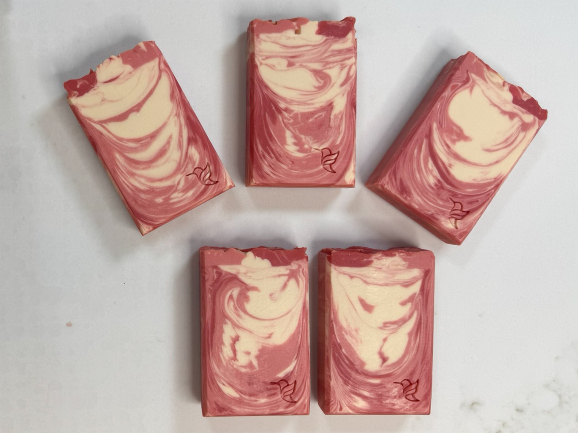 Five artisan Bloom and Amber soap bars arranged on a white quartz studio slab, showcasing unique pink and cream swirl patterns and hummingbird logo stamp.