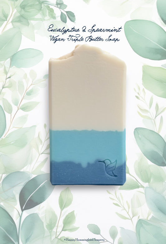 Eucalyptus & Spearmint Vegan Triple Butter Soap bar by Happy Hummingbird Soapery, featuring a white-to-blue color gradient with an embossed hummingbird logo, set against a white background with green botanical leaf illustrations.
