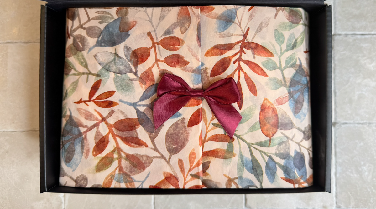 Inside view of the open black gift box displaying autumn leaf tissue paper closed with a red satin bow.
