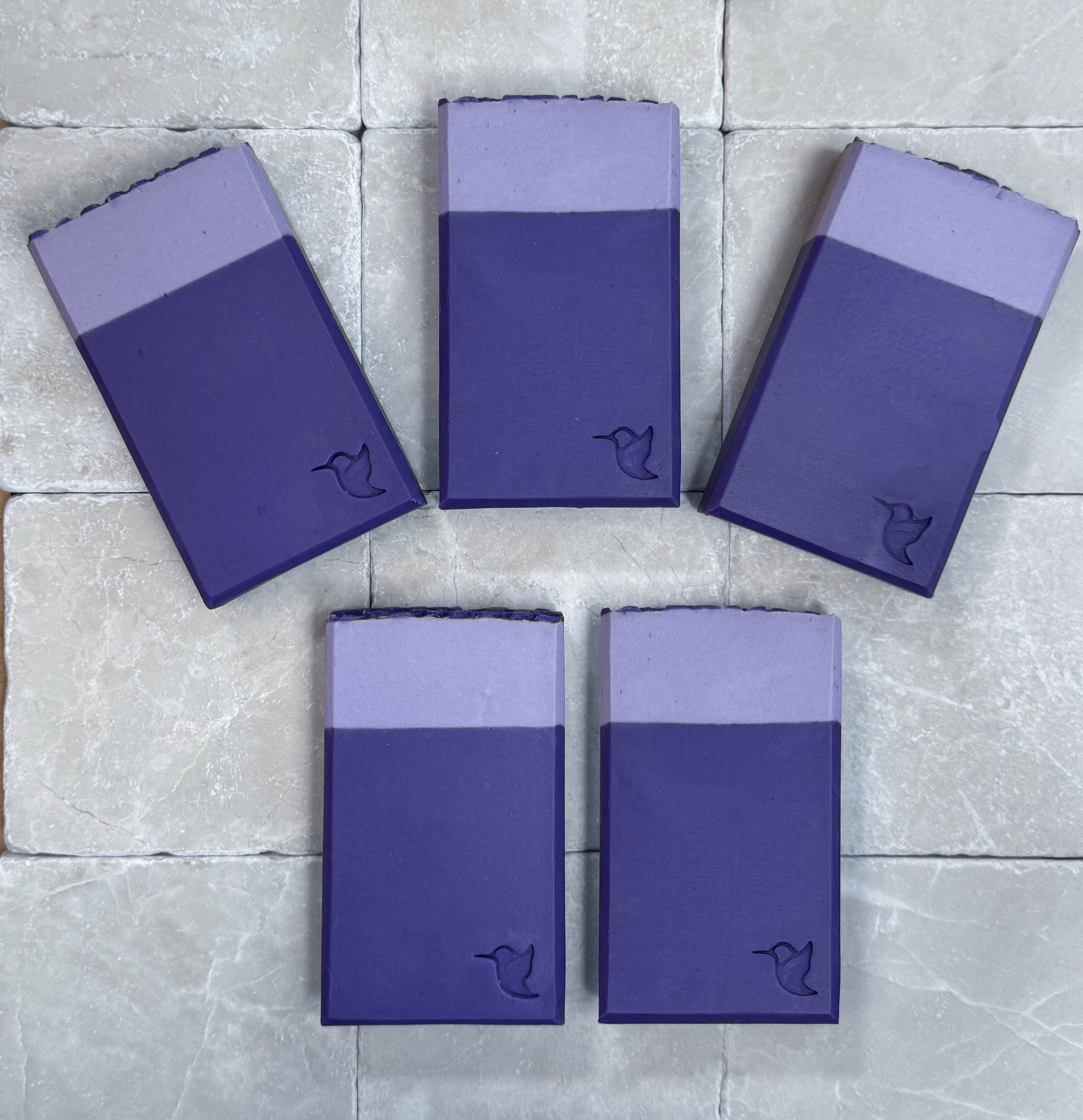 Multiple handcrafted Lavender Evening vegan soap bars by Happy Hummingbird Soapery arranged in a fan shape on a stone surface, highlighting their dual-toned purple layers and signature humming bird stamps.