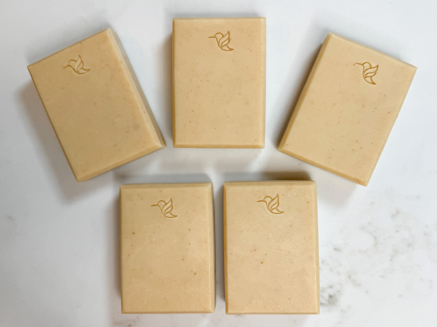 Five rectangular bars of natural beige soap arranged in a loose circle on a white quartz surface, each embossed with a small bird logo.