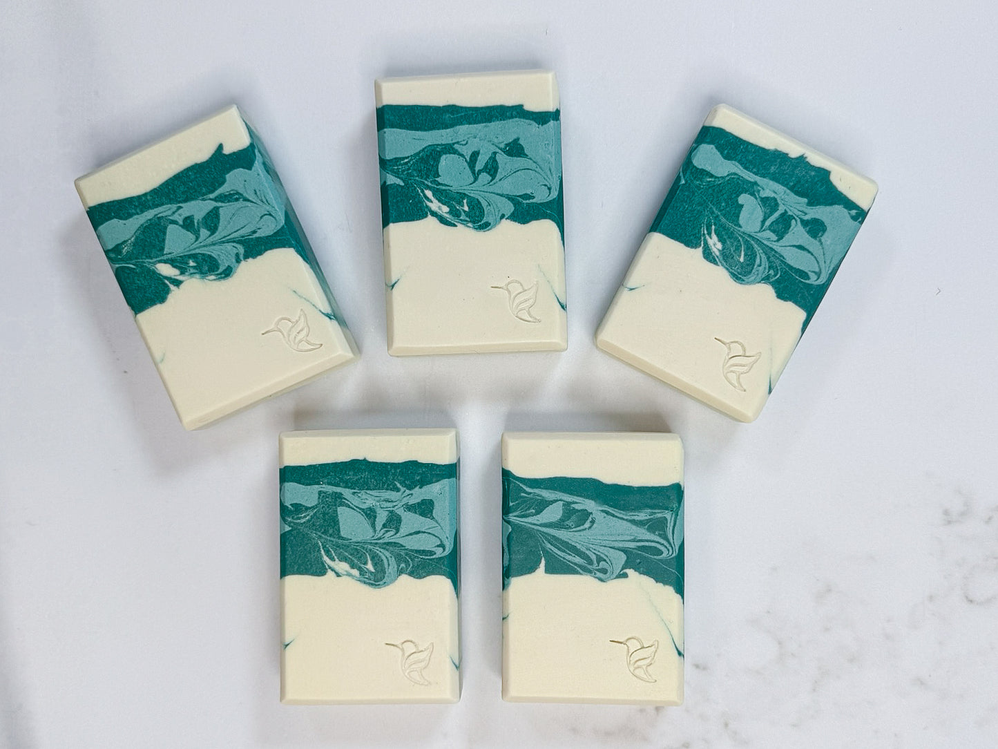 Five handcrafted bars of Wild Sage and Cucumber cold process soap arranged in a fan on a white marble background, displaying the natural variations of the deep teal and sage green marbled swirls and stamped hummingbird logos.